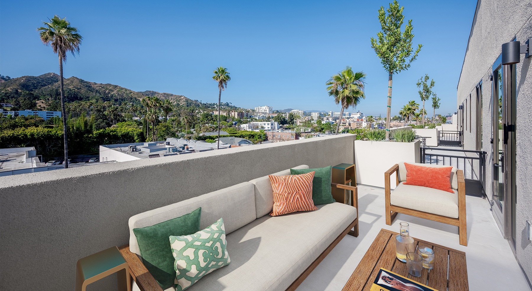 Picture of an outdoor balcony with lounge seating and palm trees and mountains in the background.